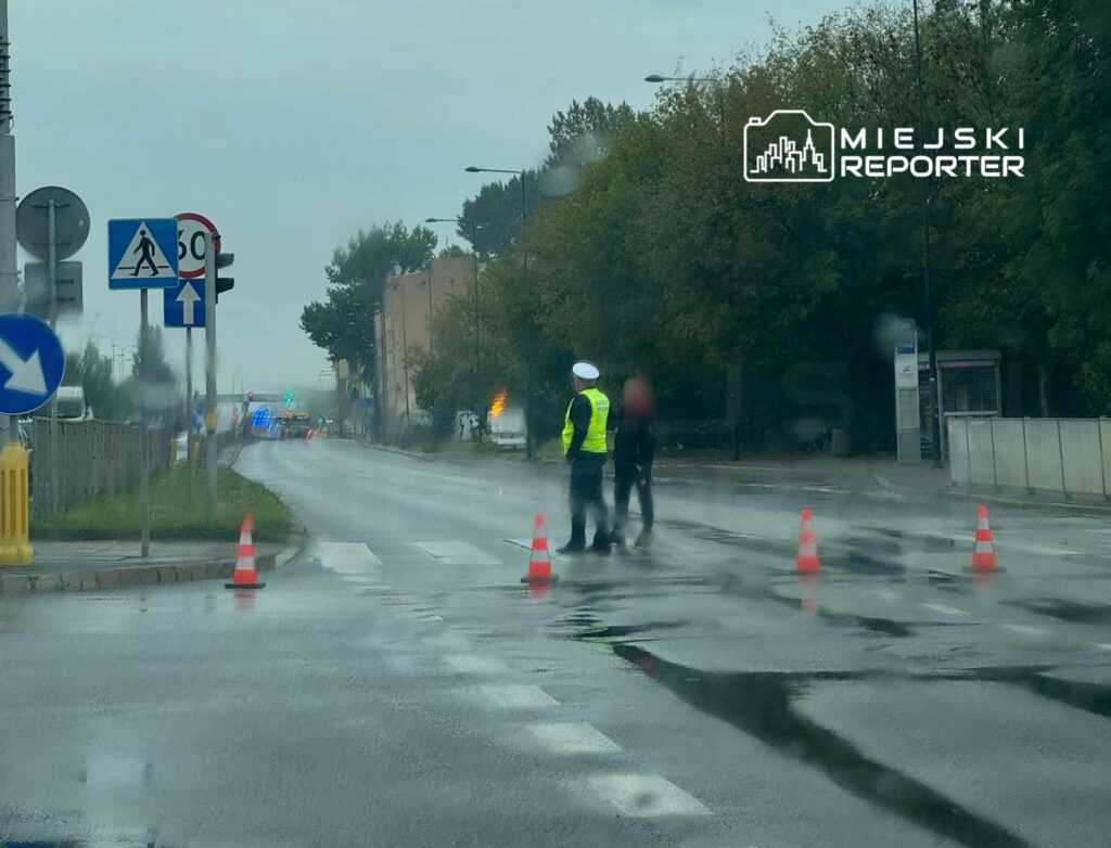 Policjant w kamizelce odblaskowej kieruje ruchem na deszczowej ulicy, obok stoi osoba.