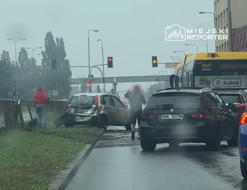 Wypadek drogowy z udziałem uszkodzonego samochodu osobowego na poboczu, obok stojących pojazdów i sygnalizacji świetlnej.