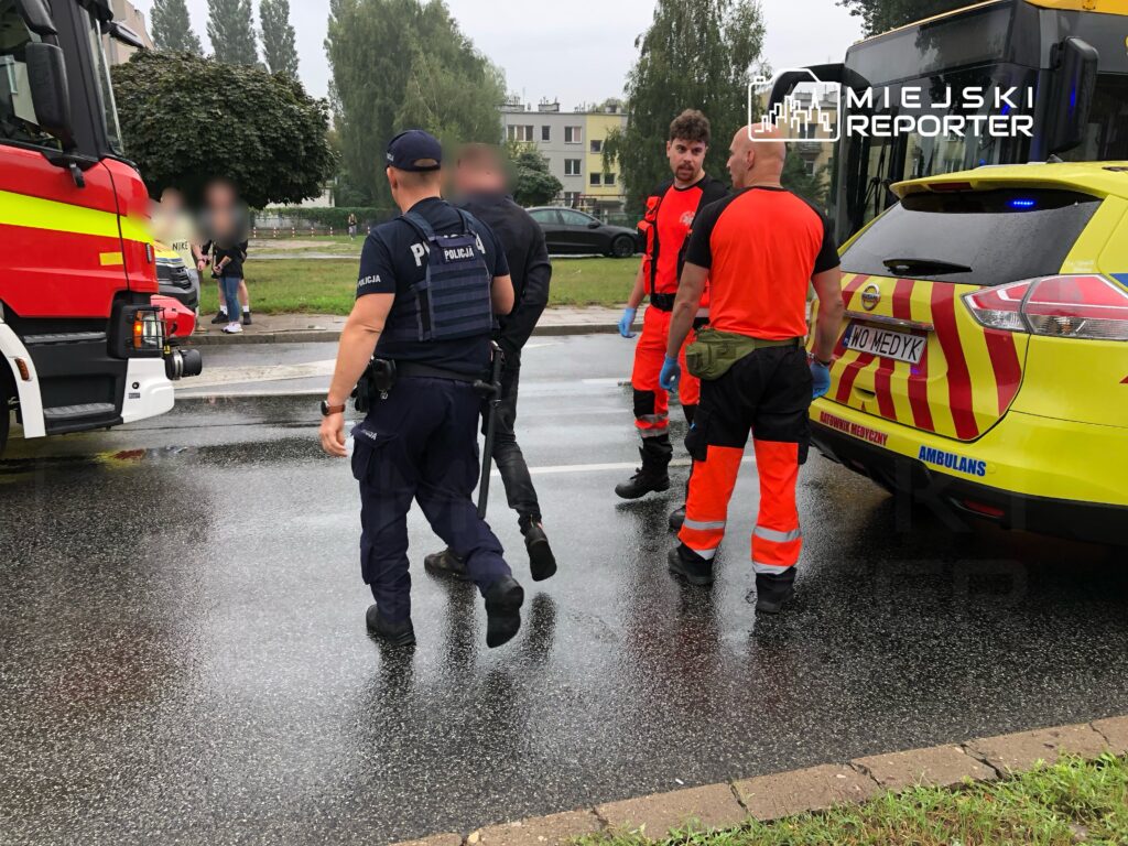 Grupa ratowników i policjantów w odzieży roboczej omawia sytuację obok wozu strażackiego i karetki na mokrej nawierzchni.