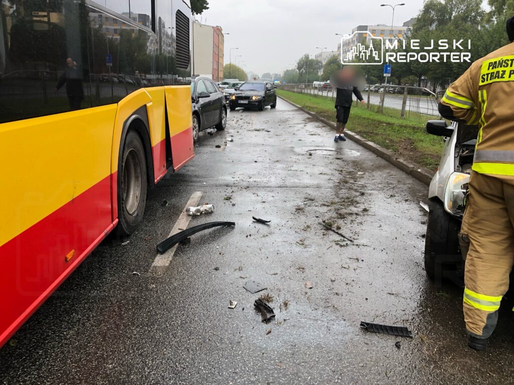Zderzenie autobusu z samochodem osobowym na drodze, wokół rozrzucone resztki i zniszczone elementy. W tle strażak bada sytuację.