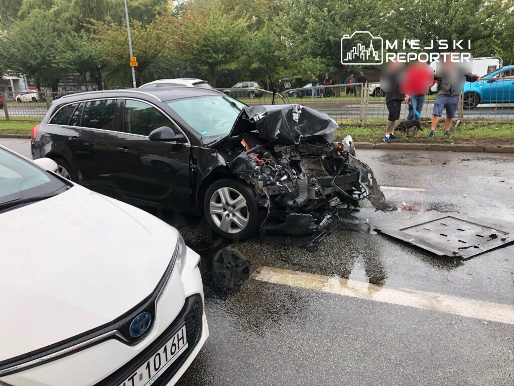Zniszczony samochód osobowy z uszkodzoną przednią częścią stoi na mokrej jezdni, obok grupy ludzi obserwujących zdarzenie.