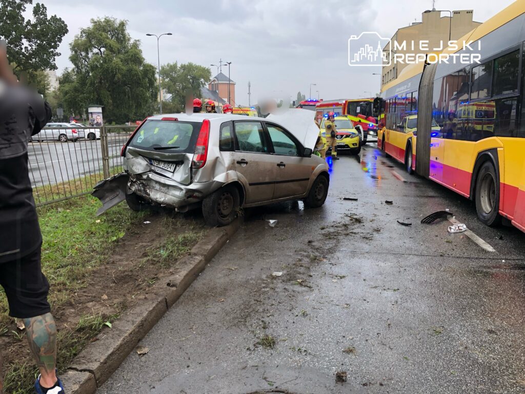 Zniszczony samochód osobowy stoi na poboczu drogi po kolizji, obok pojazdy służb ratunkowych i autobusy miejskie.