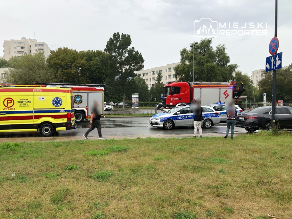 Karetka pogotowia i wóz strażacki stoją na ulicy, obok policji, w deszczowym otoczeniu osiedla mieszkaniowego.