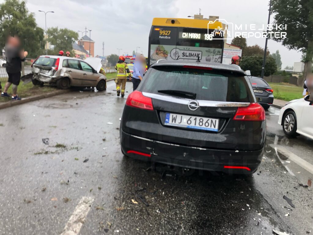 Wypadek drogowy z udziałem dwóch samochodów na deszczowej jezdni, w tle strażacy oraz autobus komunikacji miejskiej.