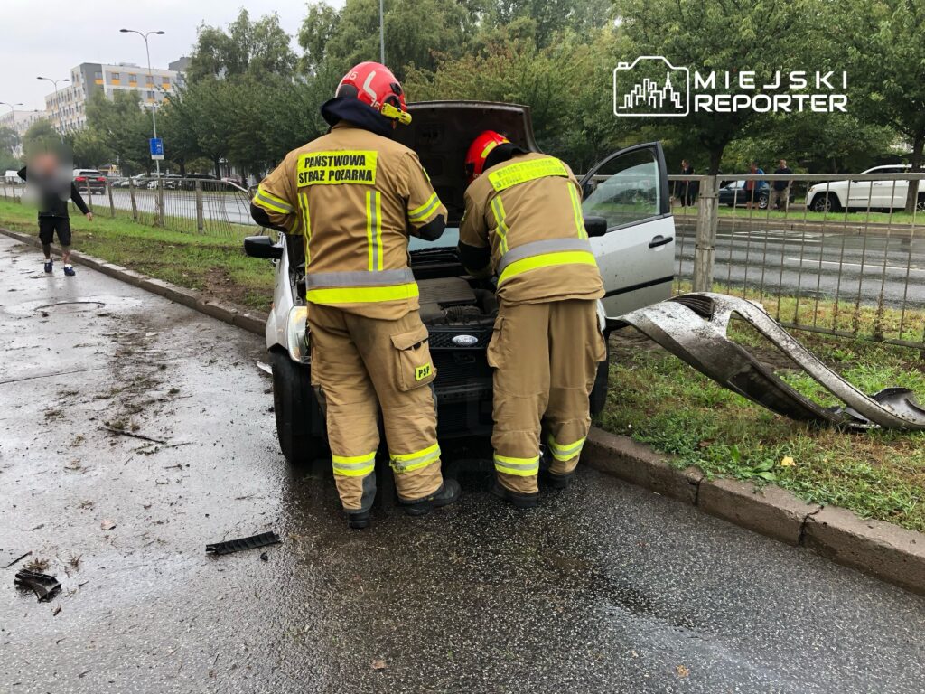 Dwaj strażacy w mundurach przeglądają silnik samochodu osobowego na poboczu drogi, obok leżą fragmenty płyty.