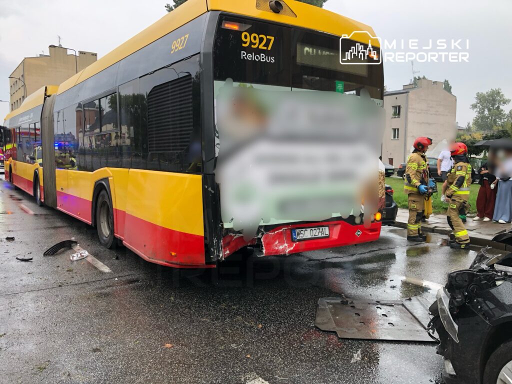 Zniszczony autobus komunikacji miejskiej na mokrej jezdni, obok strażacy rozmawiający z osobami w pobliżu.