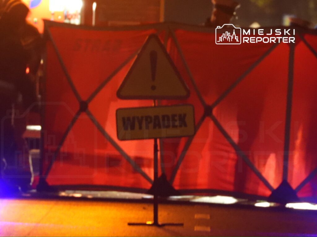 Znak ostrzegawczy "WYPADAK" ustawiony przed czerwonym parawanem, w tle widać interwencję służb ratunkowych.