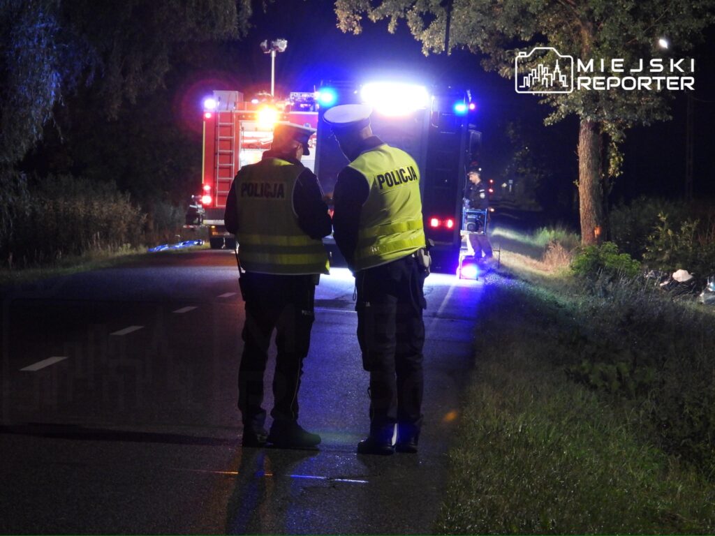 Dwóch policjantów w kamizelkach odblaskowych rozmawia na drodze, w tle widoczne pojazdy służb ratunkowych.