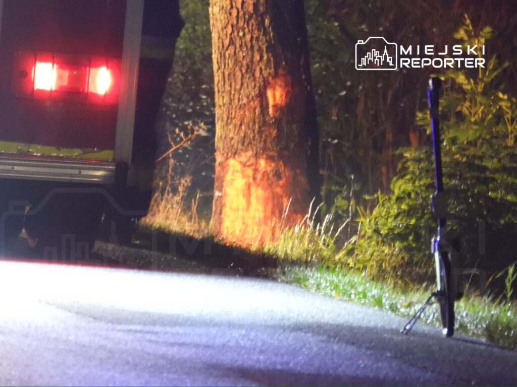 Na poboczu drogi stoi niebieska hulajnoga, a w tle widać czerwone światła wozu strażackiego oraz oświetlony pień drzewa.