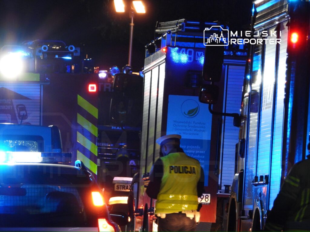 W nocy przy ulicy z widocznymi pojazdami straży pożarnej i policji, ratownicy pracują nad zabezpieczeniem terenu.
