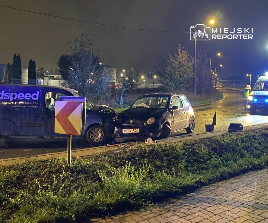 W nocy dwa uszkodzone samochody stoją na poboczu drogi, obok widoczna karetka i znak drogowy.