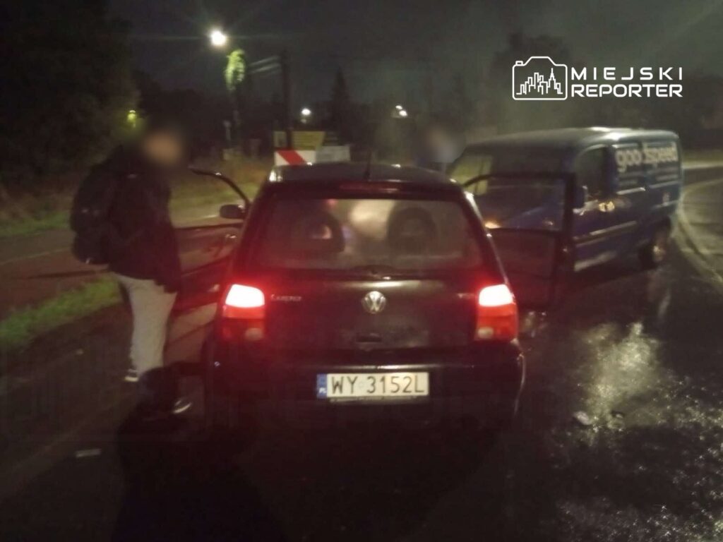 Mężczyzna w ciemnej odzieży stoi obok otwartego samochodu osobowego na mokrej drodze, obok zaparkowanego furgonu.
