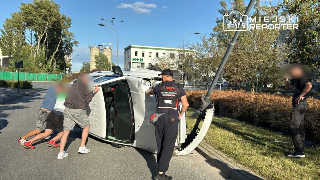 Trzech mężczyzn podnosi przewrócony samochód osobowy na ulicy, obok stoi ratownik medyczny w kamizelce.