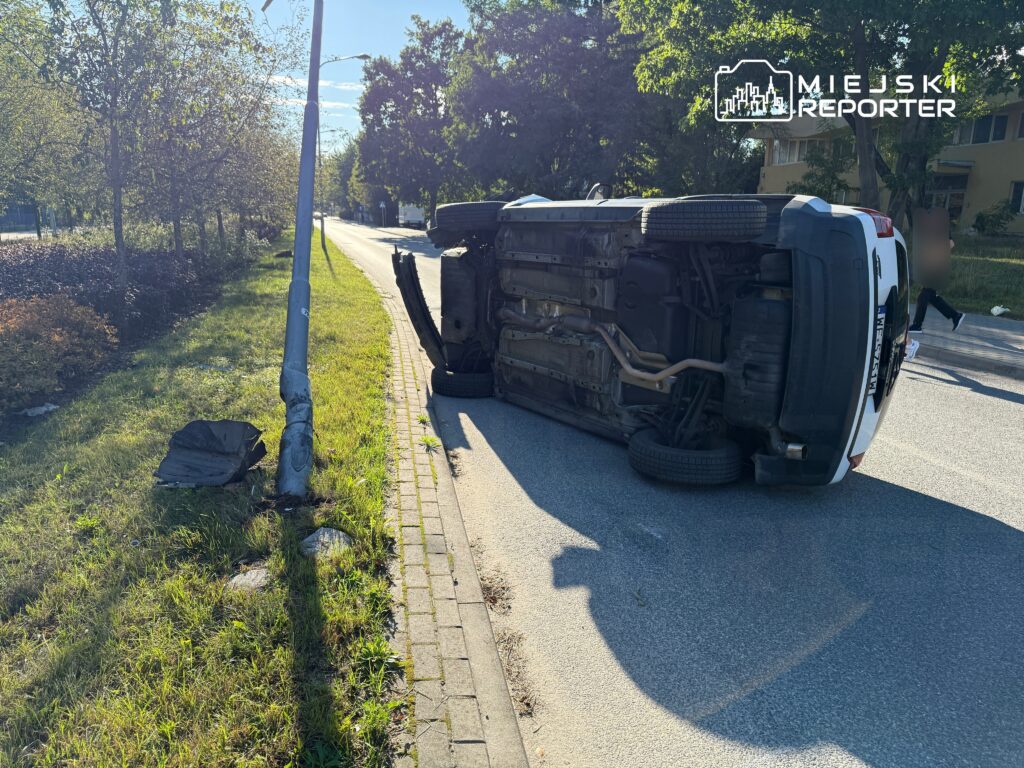 Przewrócony samochód osobowy leży na boku obok latarni, na poboczu drogi otoczonej zielenią.