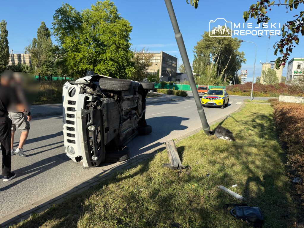 Odwrócony pojazd dostawczy leży na poboczu drogi, obok dwóch osób; w tle widać radiowóz i budynki.