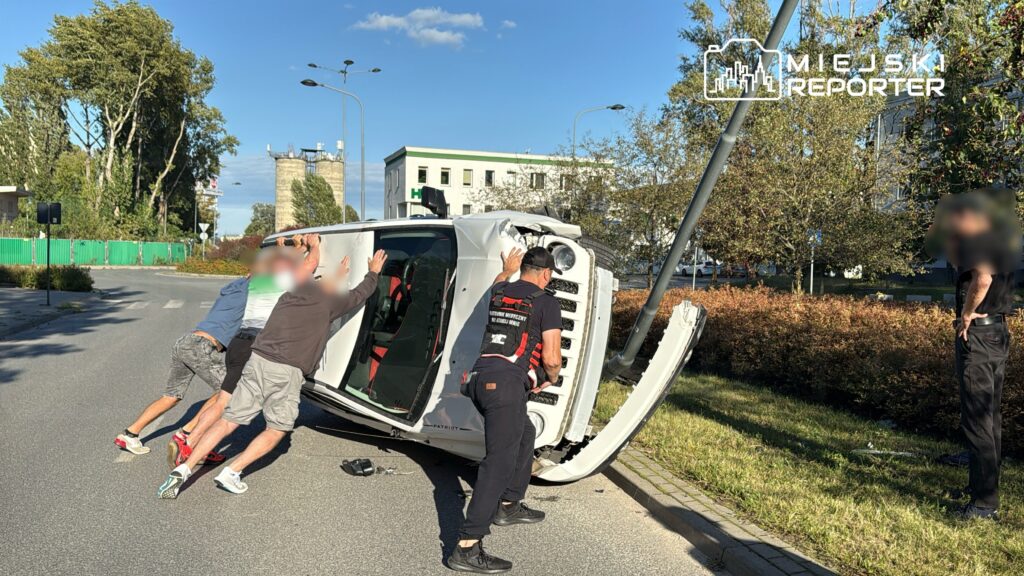 Grupa mężczyzn próbuje postawić na koła przewrócony samochód na ulicy, obok znajduje się latarnia i krzewy.