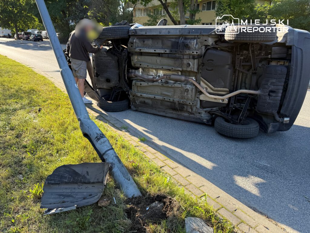 Mężczyzna w krótkich spodniach sprawdza przewrócone na bok auto obok uszkodzonego słupa.
