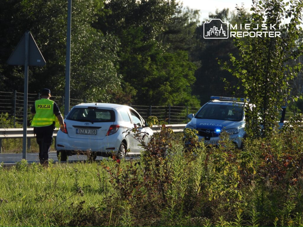Policjant w kamizelce odblaskowej podchodzi do zatrzymanego białego samochodu osobowego na drodze.