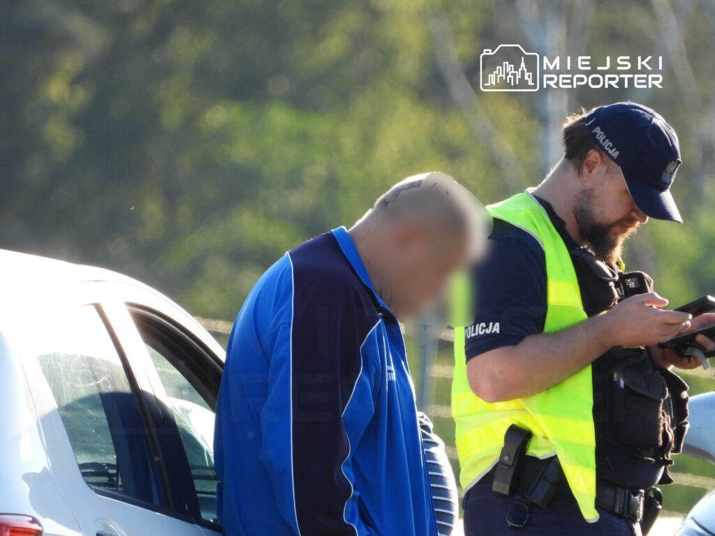 Mężczyzna w niebieskiej odzieży stoi obok białego samochodu, a policjant w żółtej kamizelce sprawdza telefon.