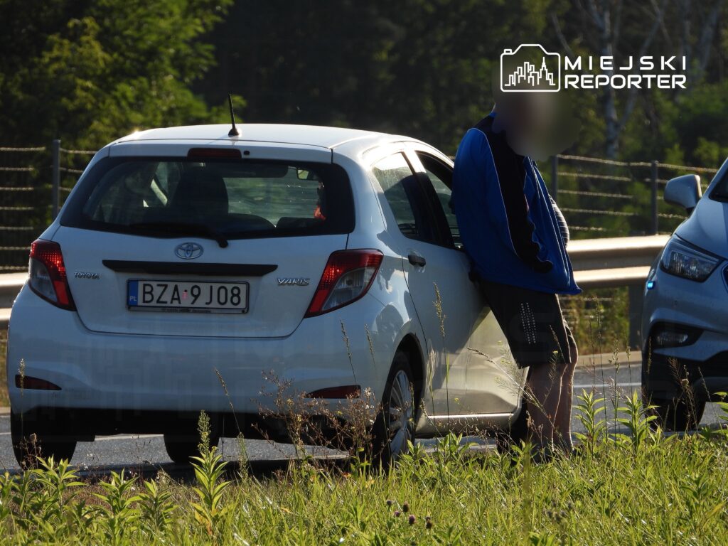 Mężczyzna w niebieskiej bluzie opiera się o białe auto na poboczu drogi, otoczenie zarośnięte trawą.