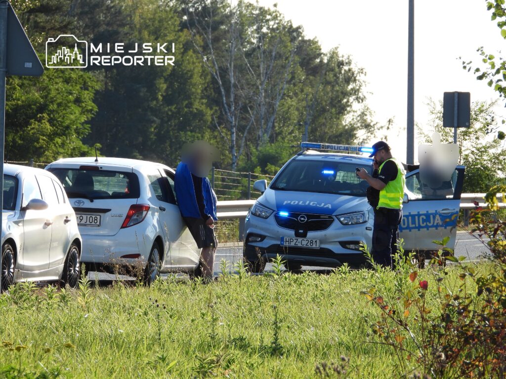 Mężczyzna w niebieskiej bluzie rozmawia z policjantem przy radiowozie na poboczu drogi.