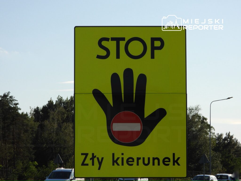 Żółty znak drogowy z symbolem ręki i napisem "STOP Zły kierunek" w otoczeniu drzew i drogi.