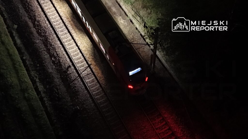 Pociąg osobowy przejeżdżający po torach oświetlonych nocnym światłem.