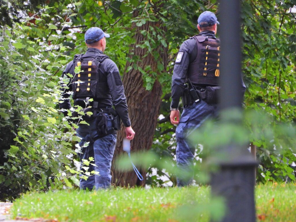 Dwóch policjantów w mundurach taktycznych spaceruje w otoczeniu zieleni, trzymając narzędzia w dłoniach.