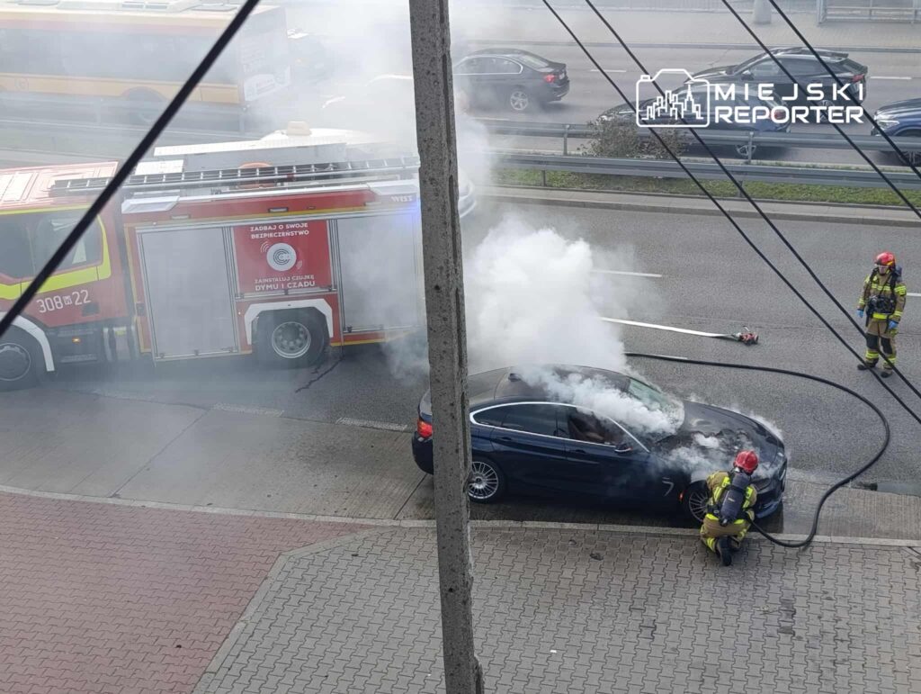 Strażacy gaszą palący się samochód osobowy na poboczu drogi, w tle widoczna strażacka ciężarówka.