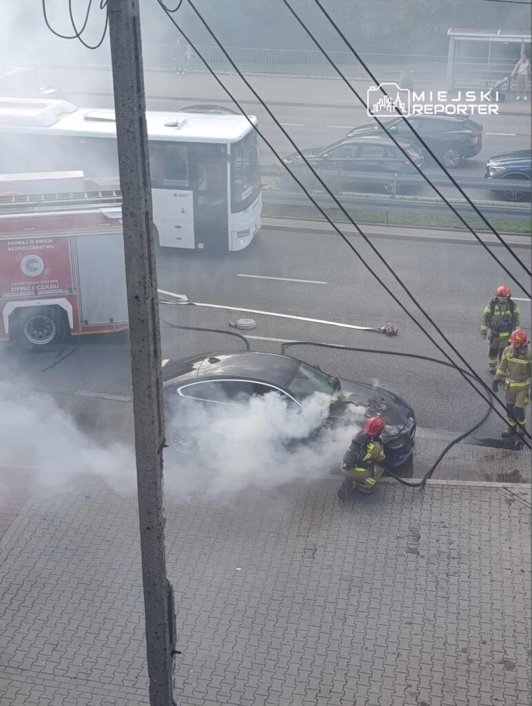 Strażak w hełmie gasi dymiący samochód na ulicy, obok stoi wóz strażacki. W tle widać przejeżdżający autobus.