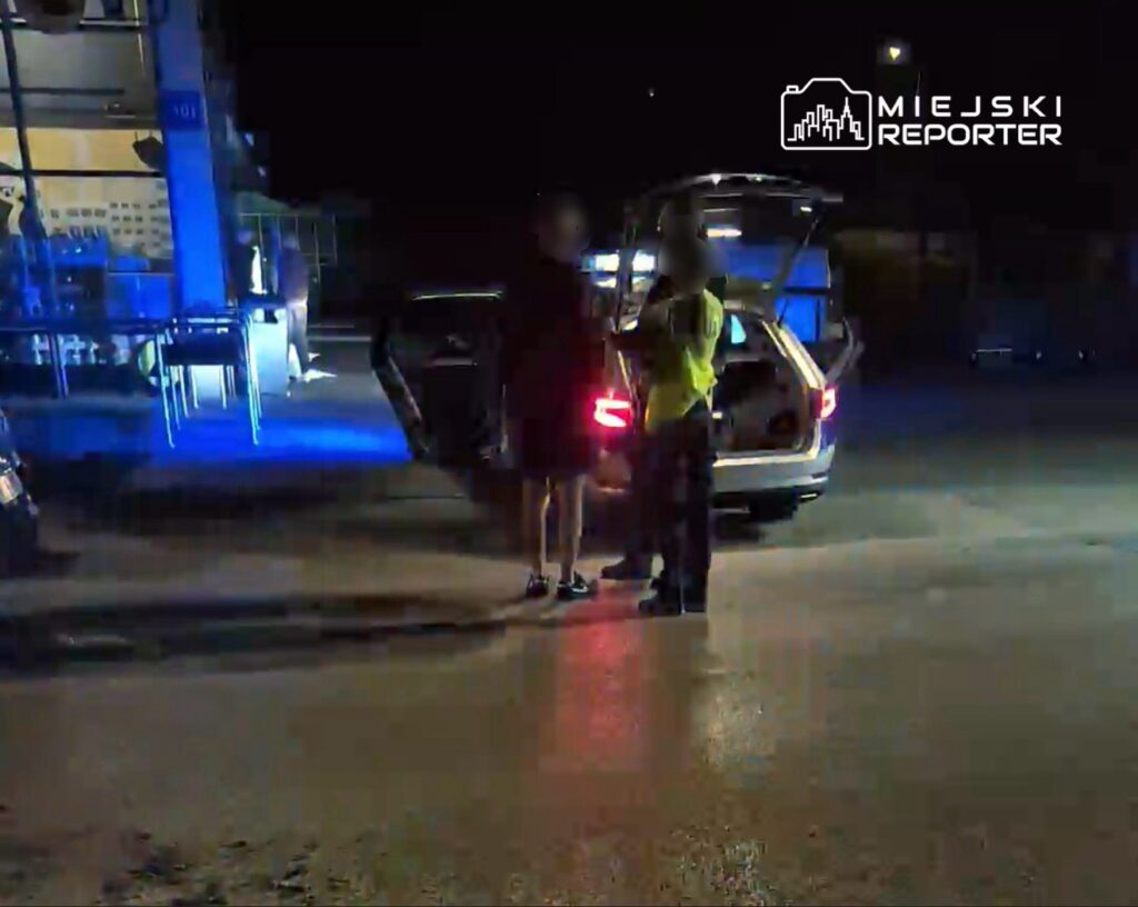 Dwaj mężczyźni rozmawiają obok otwartego bagażnika samochodu w nocy, w tle widać niebieskie światła.