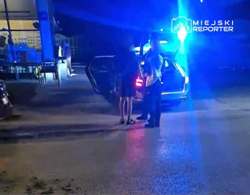 Mężczyzna w mundurze rozmawia z kobietą przy otwartym samochodzie, obok błyskają niebieskie światła.
