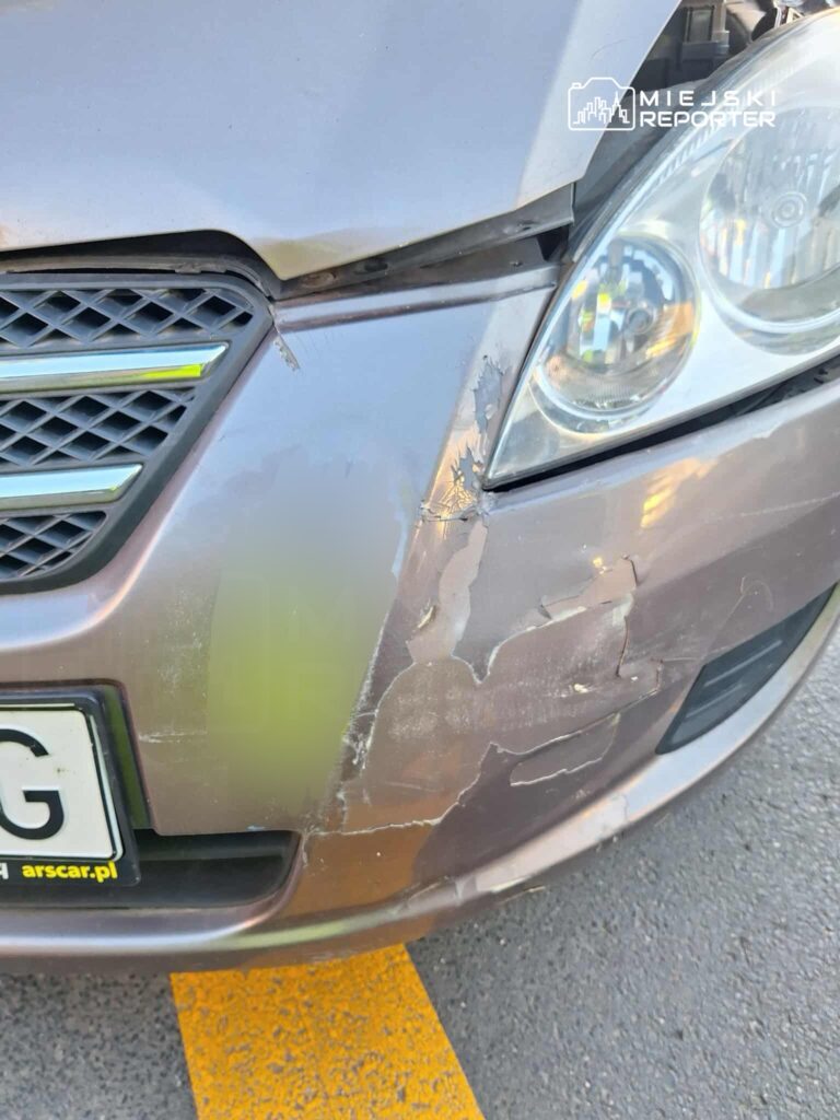 Zniszczony przód samochodu osobowego z widocznymi zarysowaniami i uszkodzeniami lakieru na parkingu.