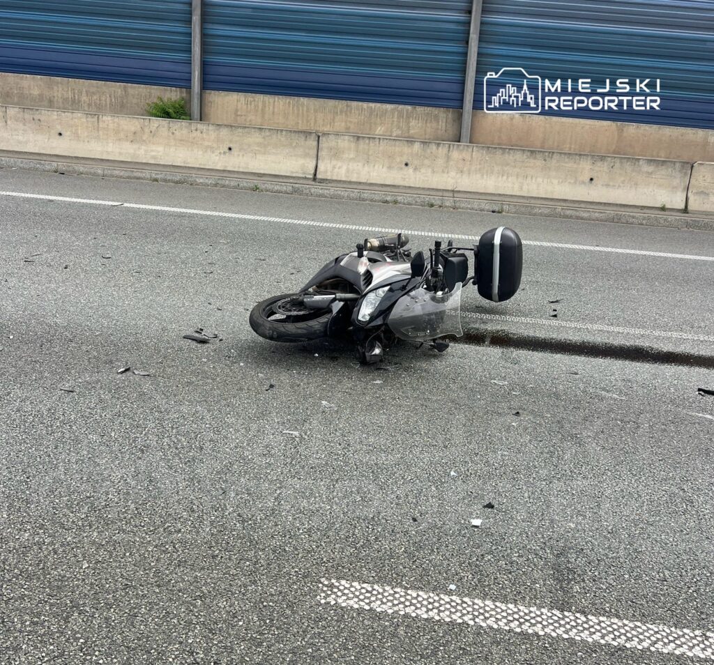 Motocykl leżący na jezdni, otoczony odłamkami, w miejskim otoczeniu.