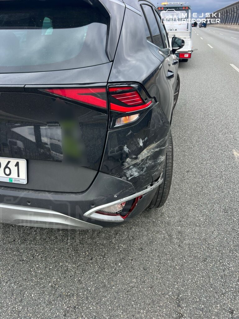 Czarny samochód osobowy z uszkodzeniami tylnym zderzaku na pasie drogowym, w tle widoczna pomoc drogowa.