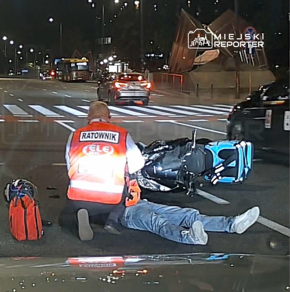 Ratownik medyczny udziela pomocy mężczyźnie leżącemu na jezdni obok przewróconego motocykla.