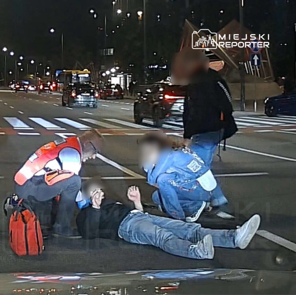 Mężczyzna leżący na jezdni, podczas gdy ratownik medyczny sprawdza jego stan, a dwie osoby obserwują sytuację.