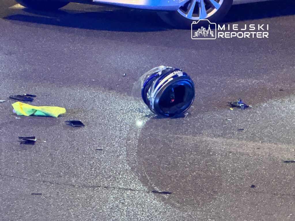 Na drodze leży rozbity kask motocyklowy oraz fragmenty szkła, obok znajduje się kolorowa chustka.