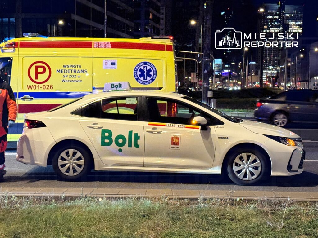 Biały samochód osobowy z logo Bolt zatrzymany obok karetki pogotowia na ruchliwej ulicy nocą.