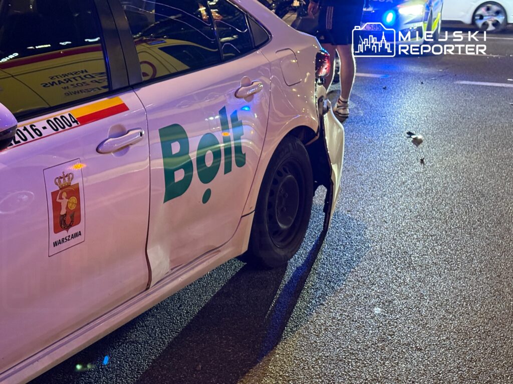 Uszkodzony samochód osobowy marki Bolt zaparkowany na ulicy, obok stojący policjant i inne pojazdy w tle.