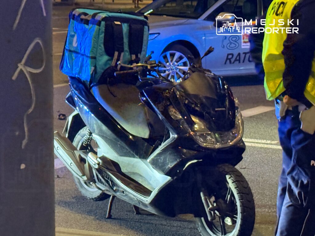 Skuter dostawczy z niebieską torbą na plecach stoi na ulicy, obok policjant w żółtej kamizelce.