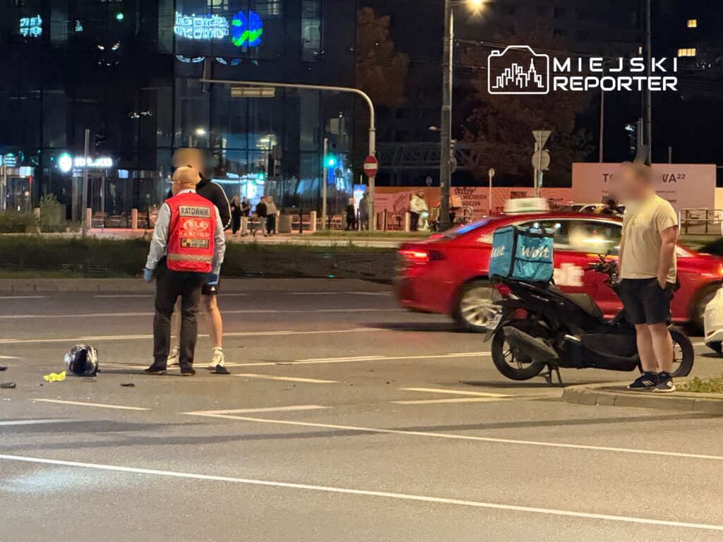 Mężczyzna w czerwonym kamizelce ratownika rozmawia z innym mężczyzną obok rozbitego skutera na ulicy nocą.