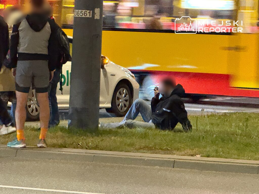 Mężczyzna w ciemnej kurtce siedzi na trawie, podczas gdy grupka osób stoi w pobliżu, a w tle przejeżdża tramwaj.