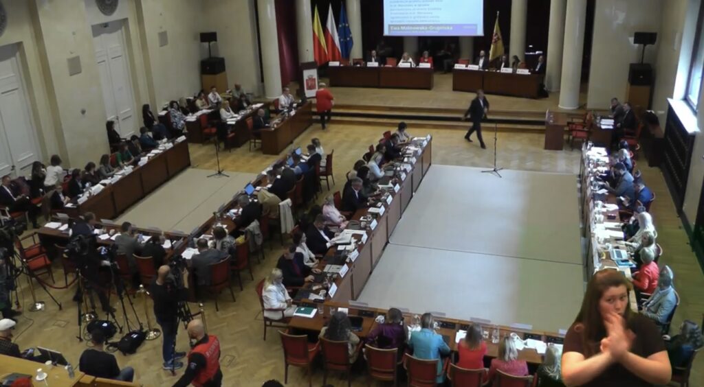 Zgromadzenie osób w sali obrad, niektórzy uczestnicy siedzą przy stołach, na ekranie prezentacja.