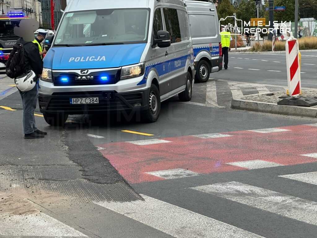 Mężczyzna w kasku rozmawia z policjantem obok radiowozu na ulicy z oznaczeniem przejścia dla pieszych.