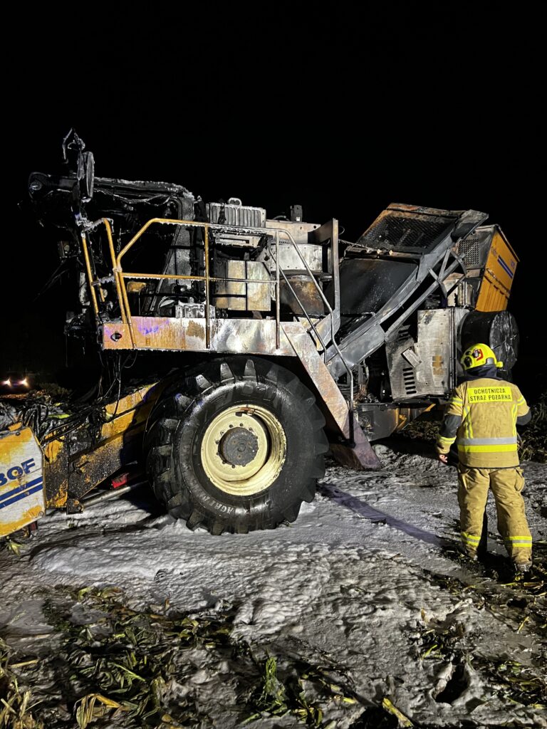 Strażak w żółtej odzieży ochronnej kontroluje spaloną maszynę rolniczą na polu w nocy. Wokół leży piana gaśnicza.