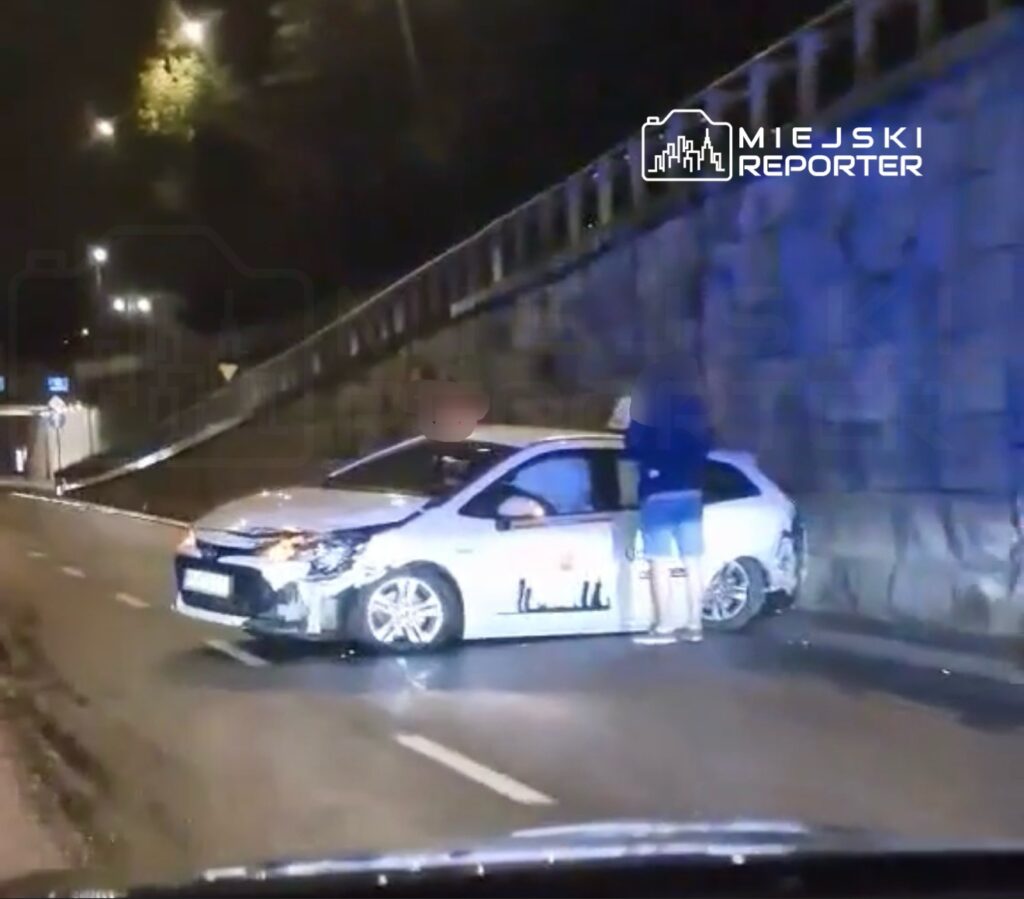 Kobieta stojąca przy uszkodzonym samochodzie na poboczu drogi nocą, w tle widoczna oświetlona jezdnia.