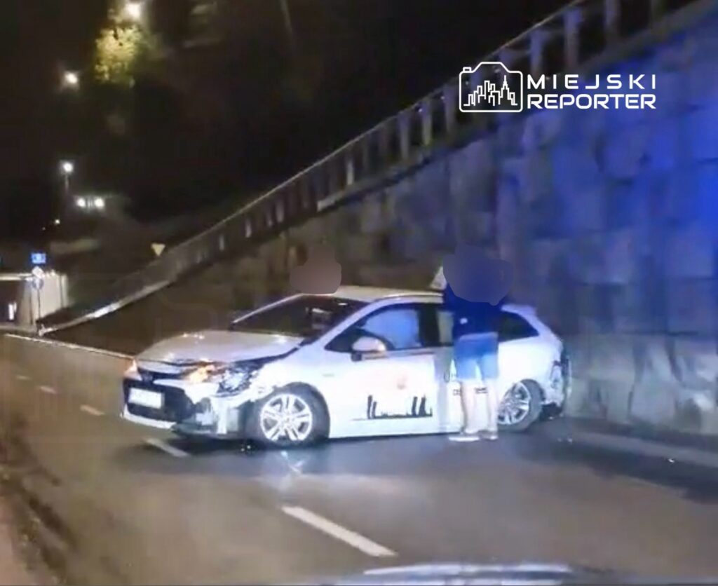 Dwie osoby stoją obok uszkodzonego samochodu na oświetlonej drodze nocą.