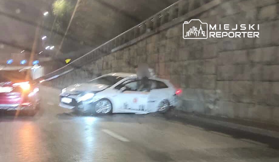 Samochód osobowy z uszkodzoną lewą stroną stoi na jezdni tunelu, obok inny pojazd. Ruch drogowy jest ograniczony.