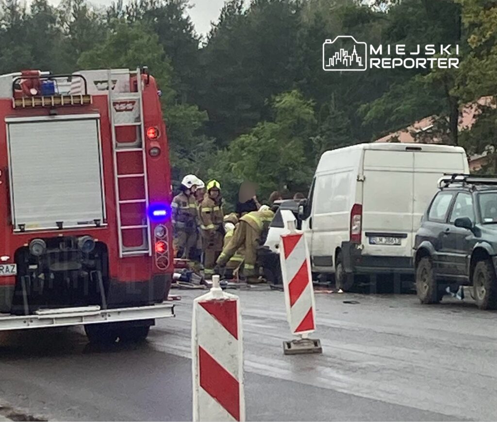 Grupa strażaków udziela pomocy poszkodowanemu na drodze, obok zaparkowanych pojazdów i oznaczeń robót drogowych.